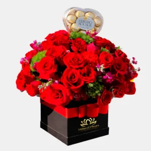 Arreglo floral de MilliersD'fleurs con abundantes rosas rojas en caja cuadrada negra con lazo rojo y estuche de chocolates Ferrero Rocher en forma de corazón.